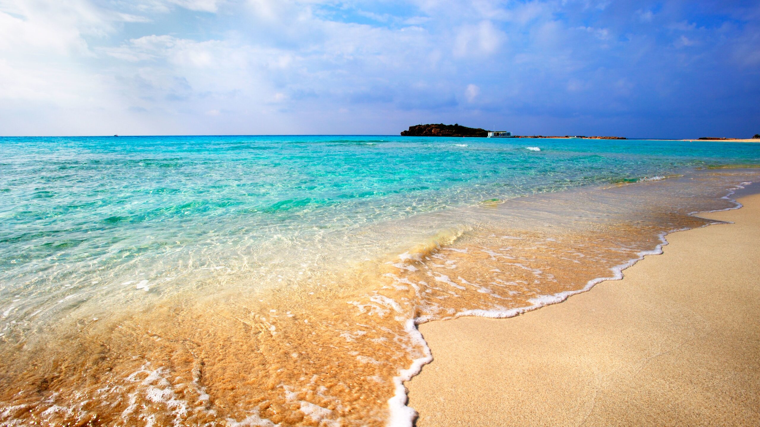 View of Nissi Beach in Cyprus with soft white sand and crystal-clear turquoise waters, showcasing the shallow shoreline and the small islet connected by a sandbar. The bright Mediterranean sun shines over the calm, inviting sea, creating a tranquil and picturesque beach scene