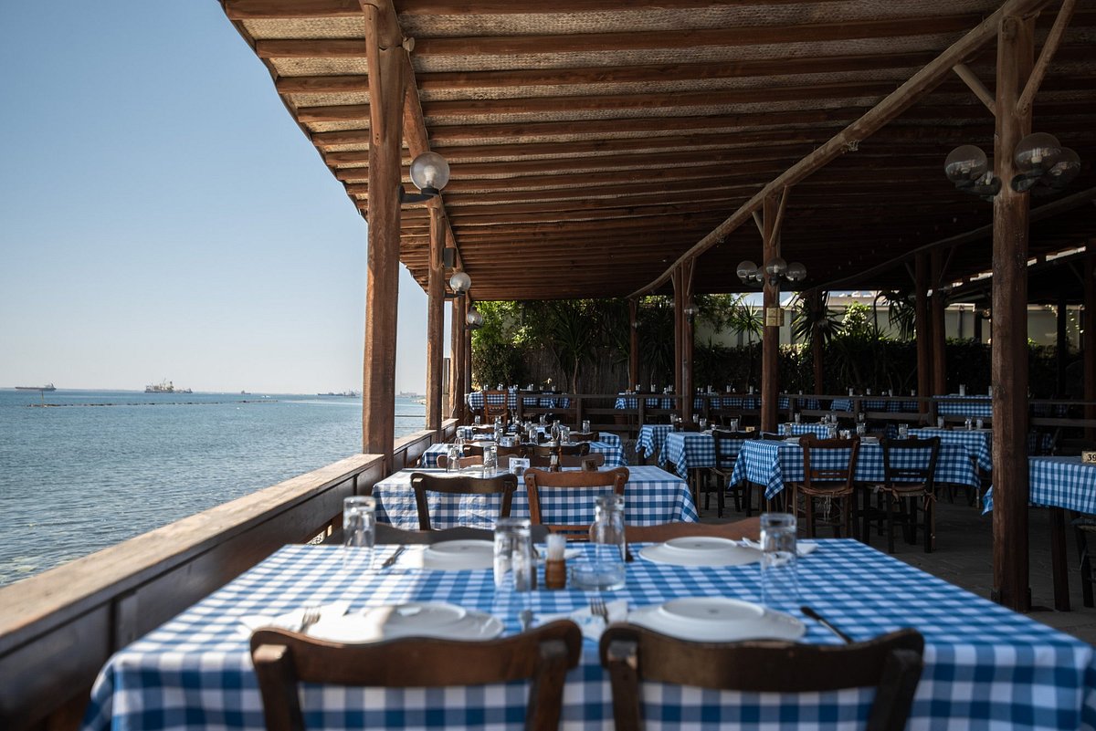 A seaside tavern with white and blue table covers, offering stunning views of the calm Mediterranean Sea. The peaceful setting features traditional Cypriot decor, with tables overlooking the serene waters, creating a relaxing and picturesque dining atmosphere